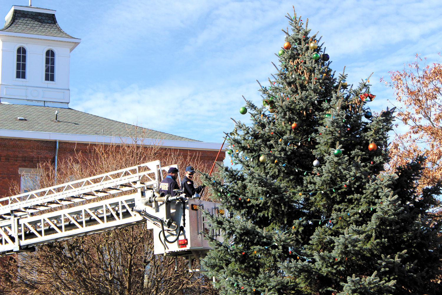 Two firepeople in a lift-truck decorate a massive pine tree with large red and green ornaments. 