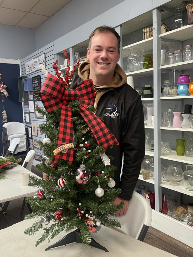 Patrick Yackmack poses with a Rotary Tabletop Christmas tree. Decorated by the high school Wrestling Squad, this tree and others will be displayed at Patrick’s shop, Crooked River Flowers, Gifts, and More on Main Street in Mantua Village.